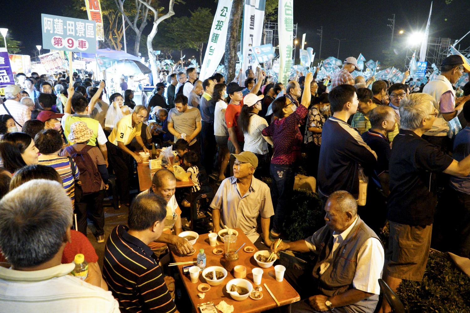陳其邁支持者在岡山造勢晚會會場外的夜市攤喝藥燉排骨,後頭的支持者則是激動的揮舞旗幟呼應舞台上的講者。 圖:張良一/攝 陳其邁支持者在岡山造勢晚會會場外的夜市攤喝藥燉排骨,後頭的支持者則是激動的揮舞旗幟呼應舞台上的講者。 圖:張良一/攝