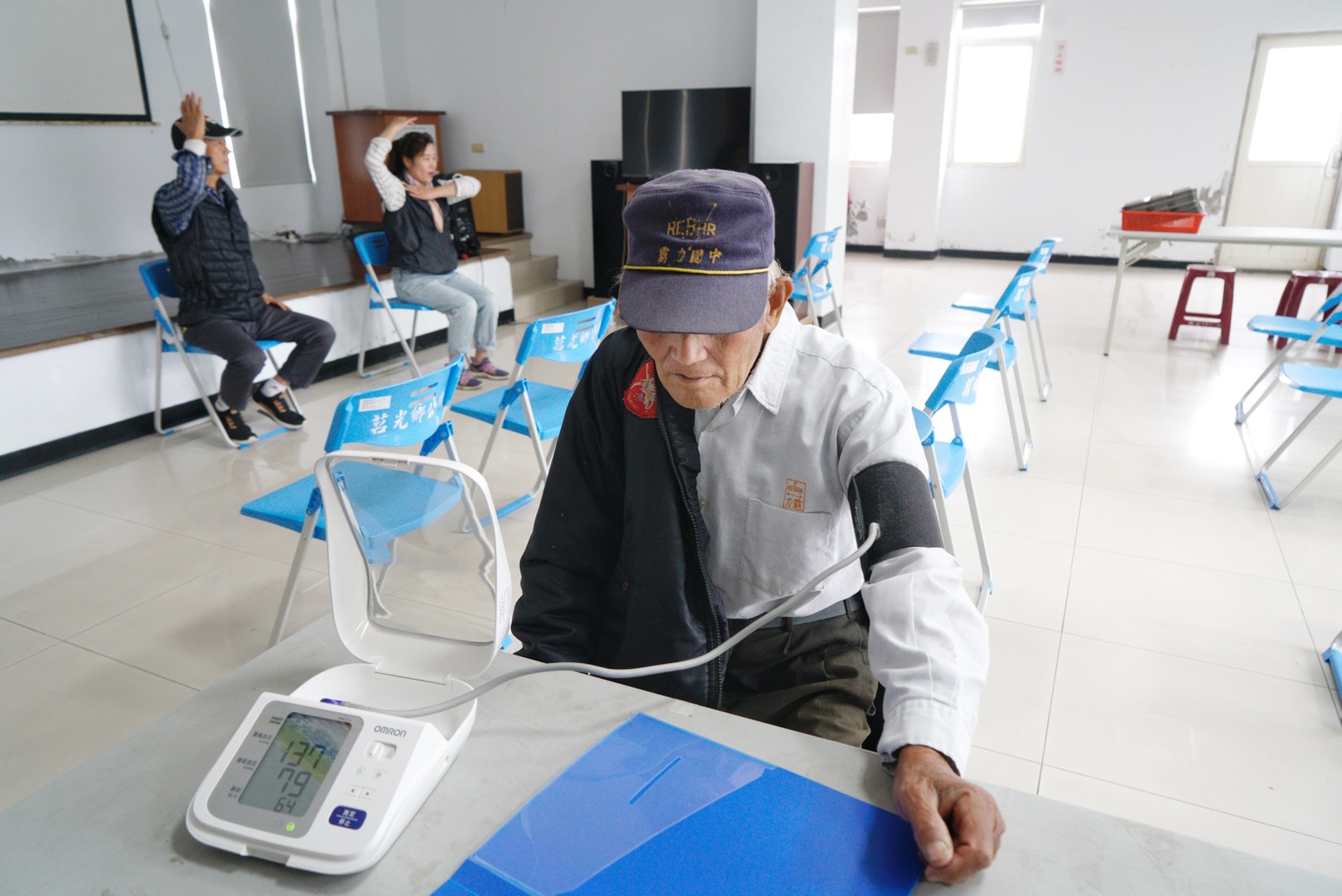 東莒一位長者出席老人共餐活動,島上衛生所的工作人員幫他量血壓,兩位居服員則正為帶動老人的肢體運動彩排。 圖:張良一/攝 東莒一位長者出席老人共餐活動,島上衛生所的工作人員幫他量血壓,兩位居服員則正為帶動老人的肢體運動彩排。 圖:張良一/攝