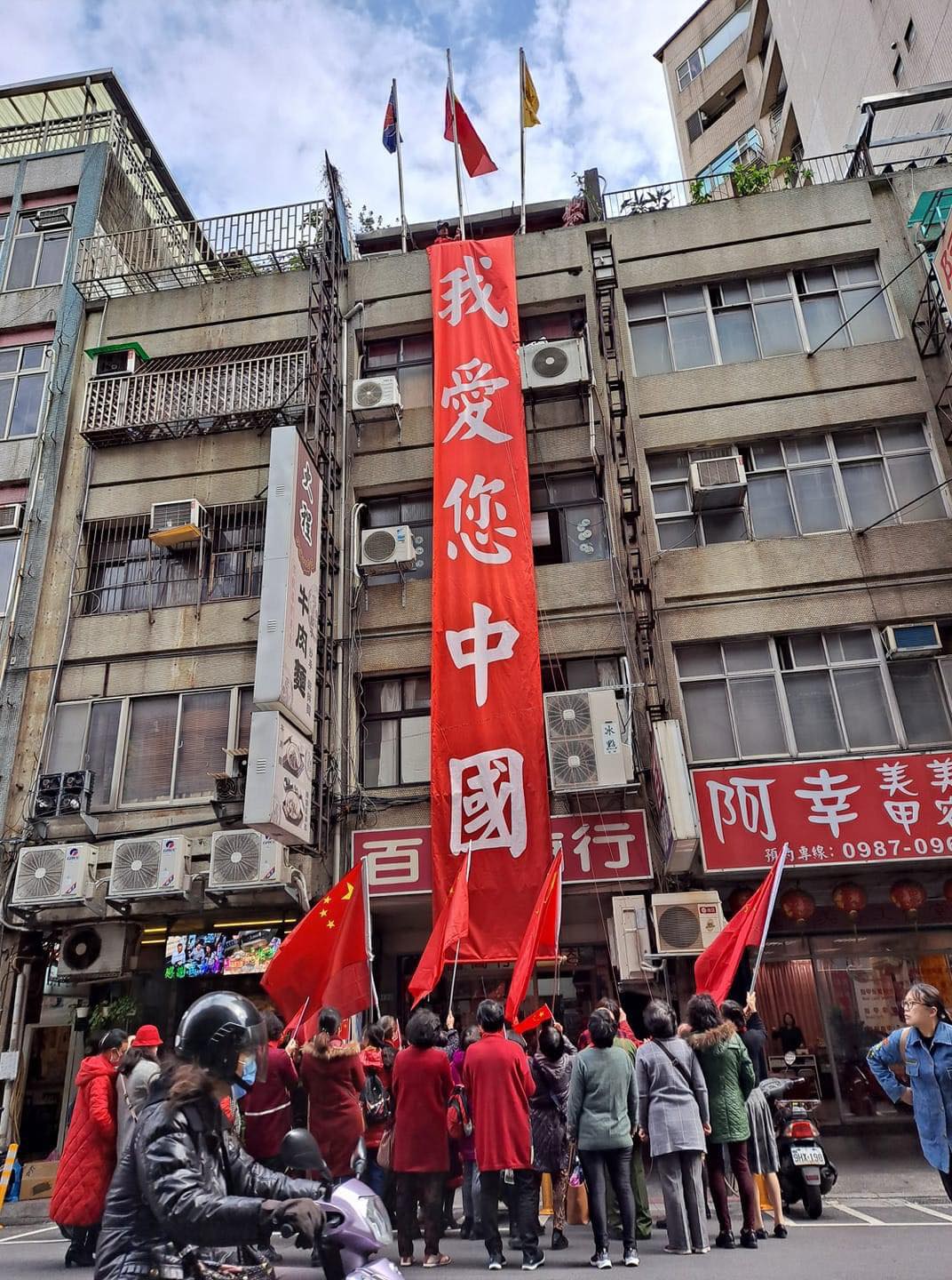 台北市街頭某處大樓外牆，卻掛上大大的紅布條，上頭寫著「我愛您中國」。1樓還有一群人高舉著「中國五星旗」   圖：翻攝自 鄉民挺起來 臉書